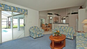 A living room with a blue carpet, two floral-patterned sofas, a coffee table, and a glass door leading to a patio with a pool. - Greater Groves 2124 (Clermont)