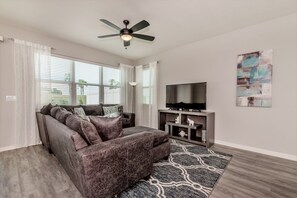 A living room with a dark brown sofa, a flat-screen TV on a wooden stand, a ceiling fan, and a patterned rug. - Compass Bay 5153 (Kissimmee)