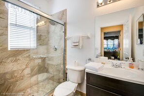 A bathroom with a glass-enclosed shower, a toilet, a sink with toiletries, and a mirror reflecting a cityscape. - Highlands Reserve Golf & Country Club 215 (Davenport)