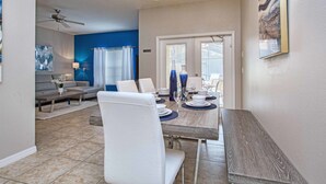 Un coin repas avec une table en bois dressée pour quatre personnes, des chaises blanches et un ventilateur de plafond.
