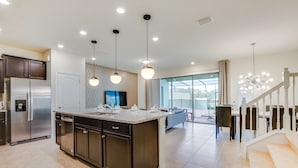 Una cucina moderna con una grande isola, elettrodomestici in acciaio inossidabile, illuminazione a sospensione e vista su un'area pranzo con una scala.