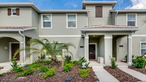 A townhouse with a light gray exterior, white trim, and a tiled roof. There are two visible doors, one with a small porch area, and a number '4958' on the wall. - Storey Lake 4958 (Kissimmee)