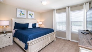A bedroom with a bed, bedside tables, a desk, and a window with blinds. - Windsor at Westside 1852 (Kissimmee)