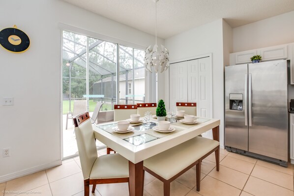 A modern kitchen with a stainless steel refrigerator, a dining table set with white plates and bowls, and a large window offering a view of the outdoors.