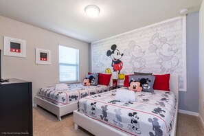 A bedroom with two beds, each with Mickey Mouse themed bedding. There are framed certificates on the wall and a window with blinds.