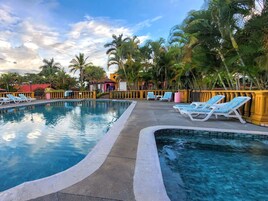Una piscina con sillones de descanso, palmeras y un edificio de techo rojo al fondo.