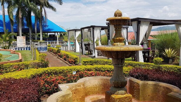 Un jardín con una fuente central, un edificio de techo azul y una zona de estar cubierta.