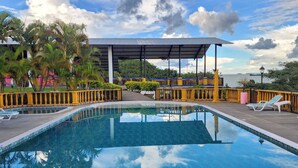 Una piscina cristalina con un área de estar cubierta y sillones de descanso, rodeada de palmeras y vista al agua.