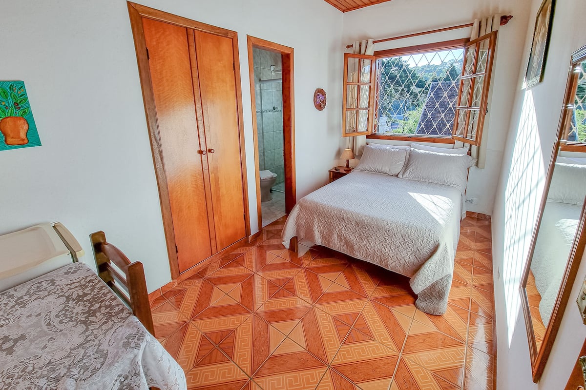 A bedroom with a bed, a wooden wardrobe, a bathroom, and a window with a view of greenery.