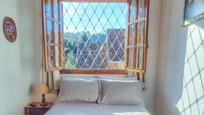 Una cama con colcha, una mesita de noche con lámpara y una ventana con vista a edificios.