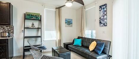 A modern living room with a black leather sofa, a metal chair, a small table, and a ceiling fan. There is a shelf with decorative items and a painting on the wall.