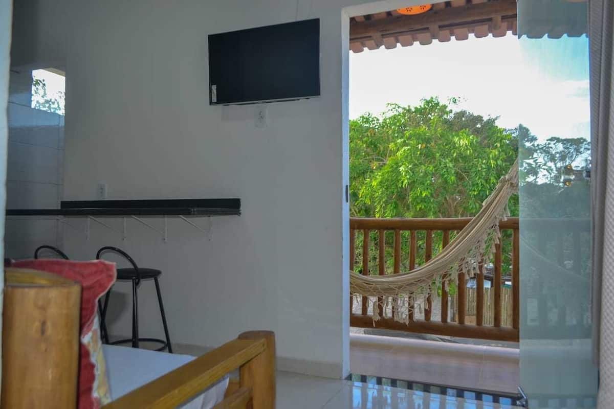 A room with a bed, a TV mounted on the wall, a hammock on the balcony, and a view of greenery outside.