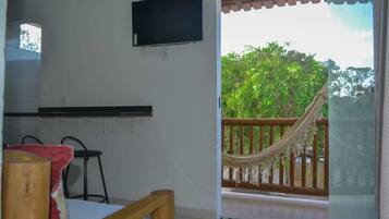 Quarto com cama, televisão na parede, rede na varanda e vista para o verde lá fora.