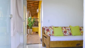 A room with a wooden sofa with floral cushions, a glass door leading to a hallway, and a potted plant. - Villa Nobre Residence (Porto Seguro)