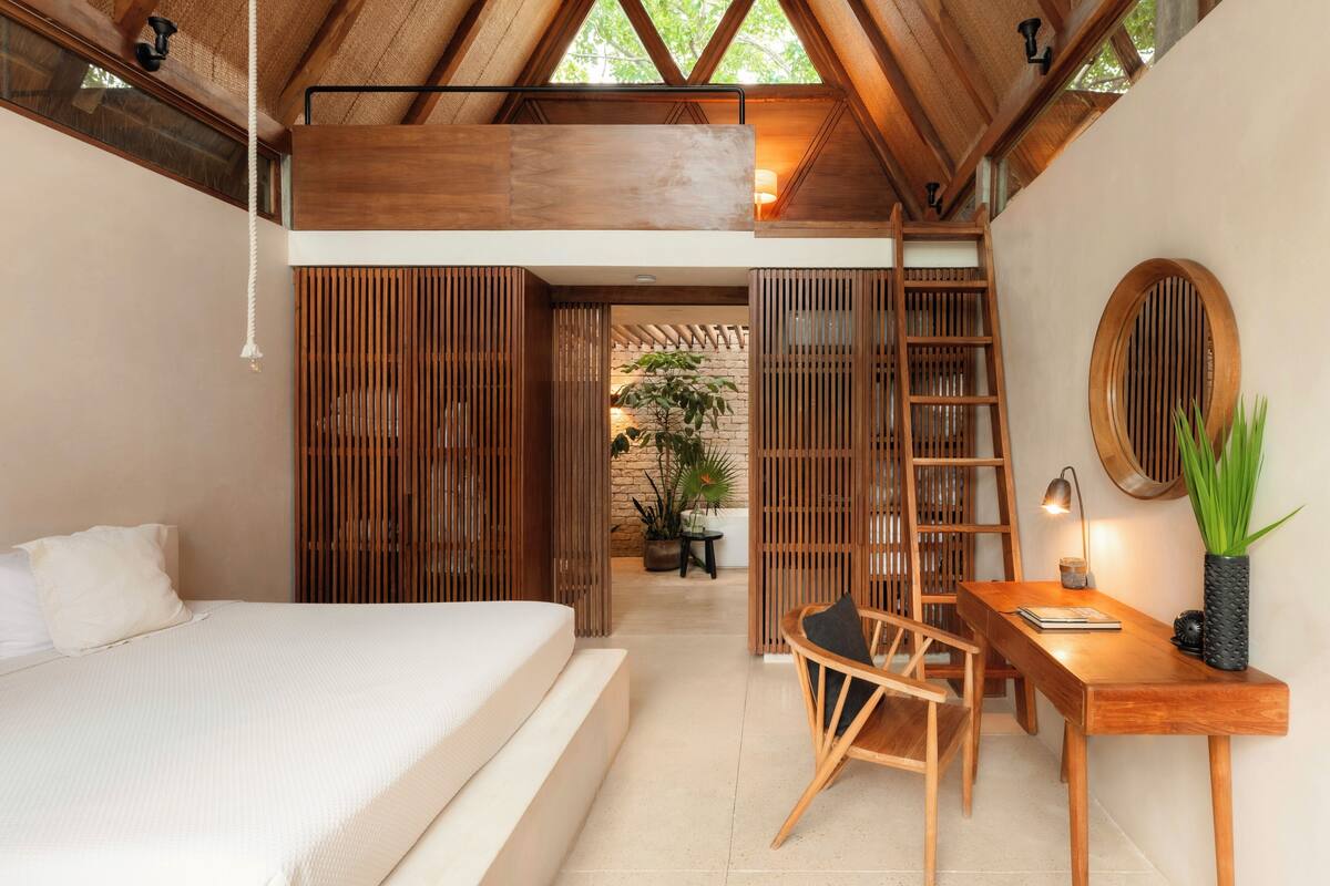 A bedroom with a bed, wooden desk, chair, and a small table with a plant.