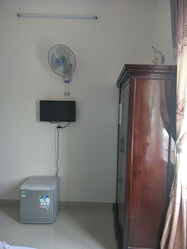 Una habitación con un armario de madera, un ventilador de pared, un televisor y un refrigerador pequeño.