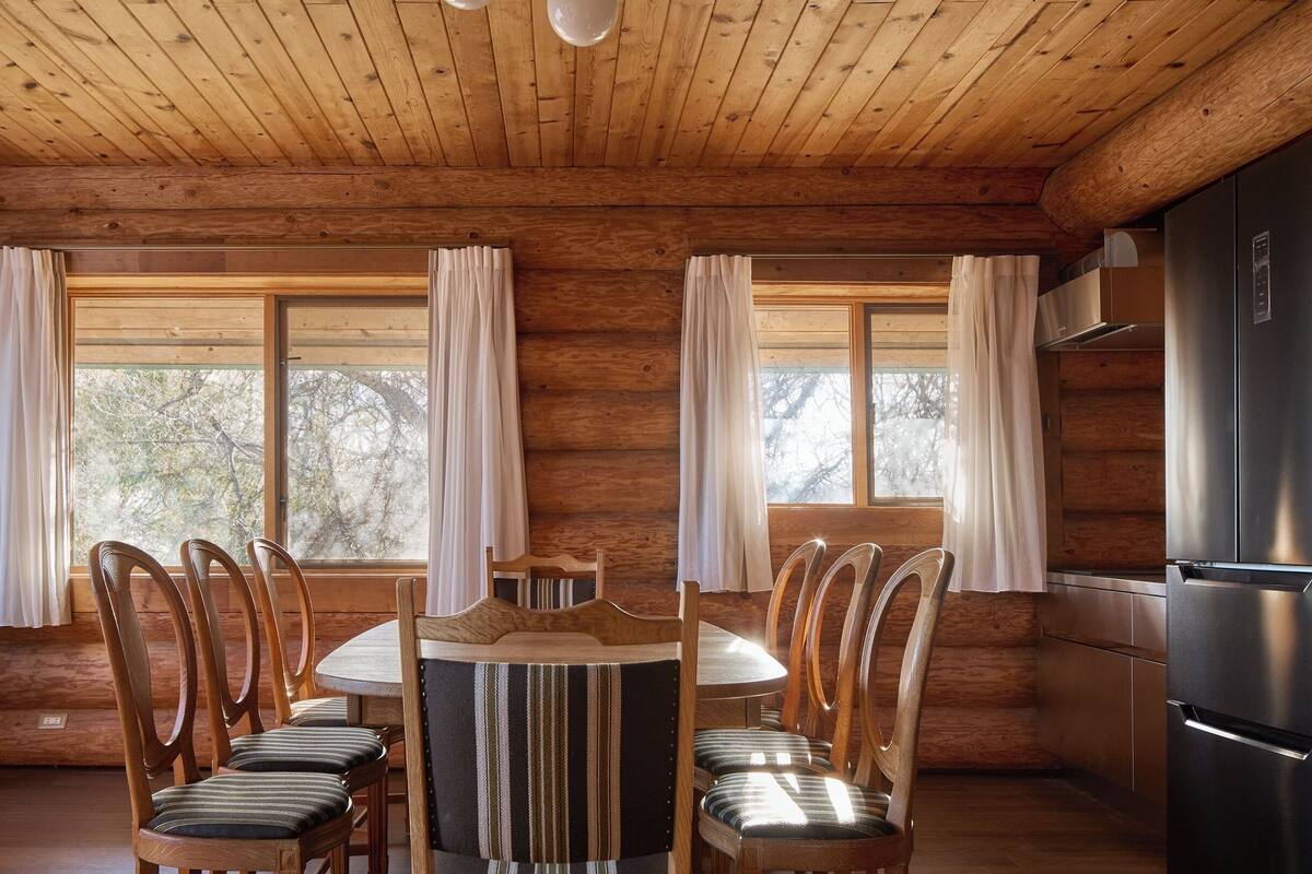 A wooden cabin interior with a dining table surrounded by chairs, large windows with curtains, and a kitchen area with a refrigerator.