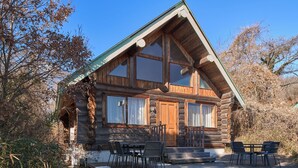 A log cabin with a steep gabled roof, large windows, and a front porch with outdoor seating. - Mission Hills Hotel (Minano)
