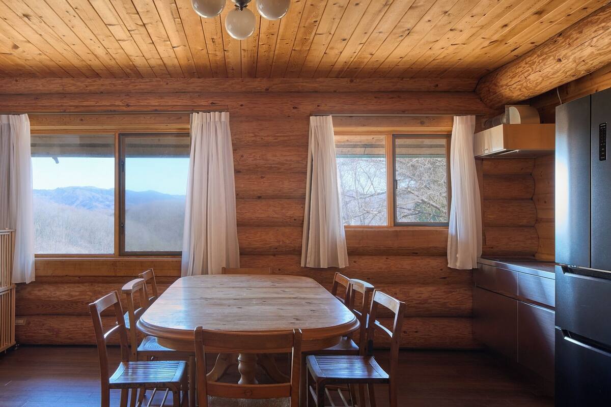 A wooden cabin interior with a round dining table, six chairs, a kitchen area with a refrigerator, and large windows offering mountain views.