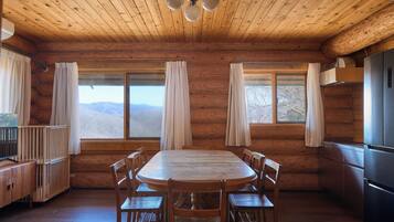 A wooden cabin interior with a round dining table, six chairs, a kitchen area with a refrigerator, and large windows offering mountain views.