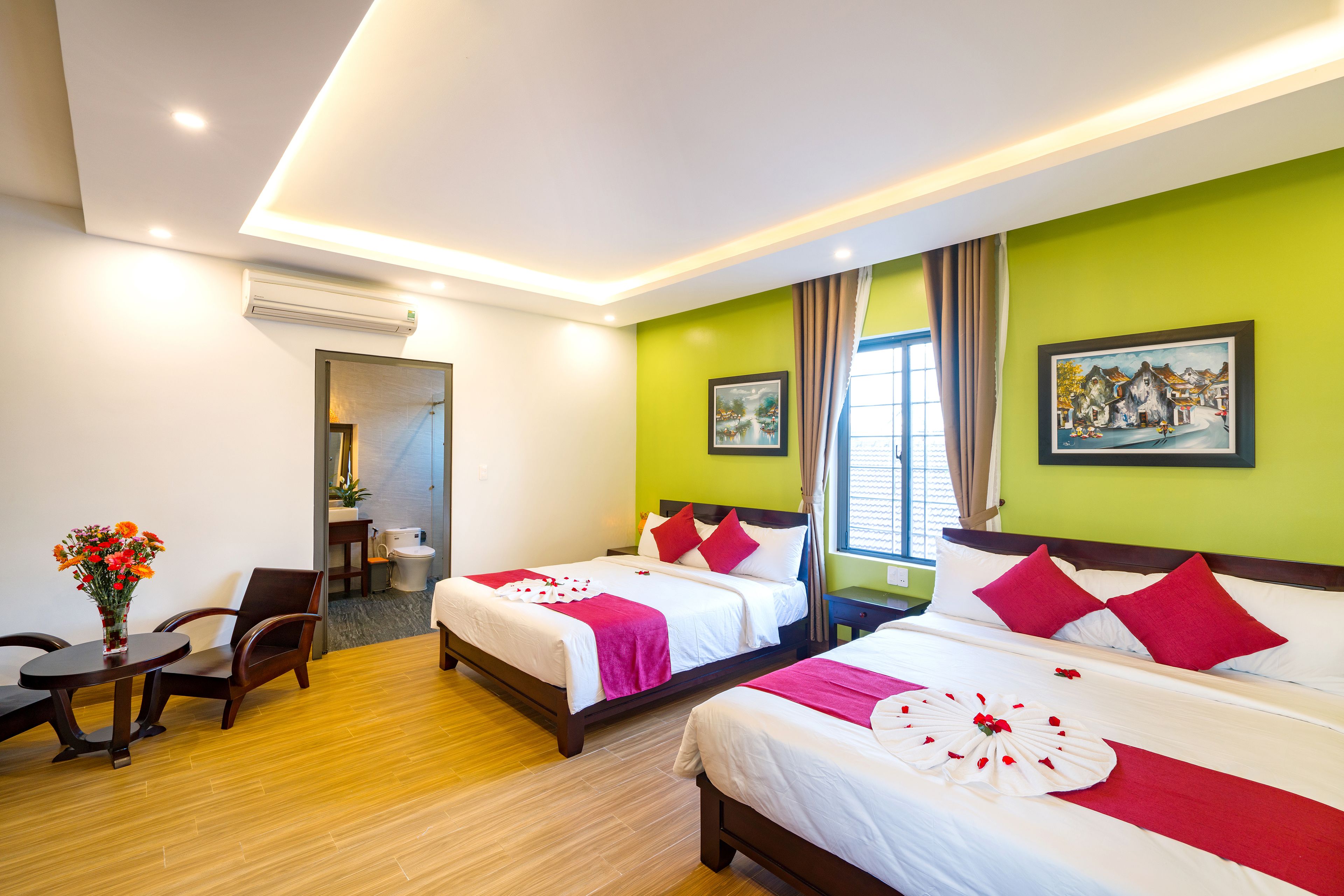 Une chambre d’hôtel avec deux lits, un coin salon avec une table et des chaises, une fenêtre avec des rideaux et des œuvres d’art accrochées aux murs.