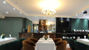 A restaurant with a central dining table, green upholstered chairs, a chandelier, and a bar area with a modern design. - UB Grand Hotel (Ulaanbaatar)