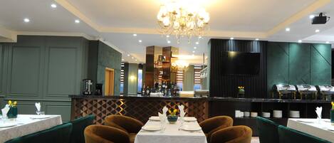 A restaurant with a central dining table, green upholstered chairs, a chandelier, and a bar area with a modern design.