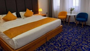 A hotel room with a large bed, two orange pillows, a bedside table with a lamp, a small round table with a chair, and a blue carpet. - UB Grand Hotel (Ulaanbaatar)