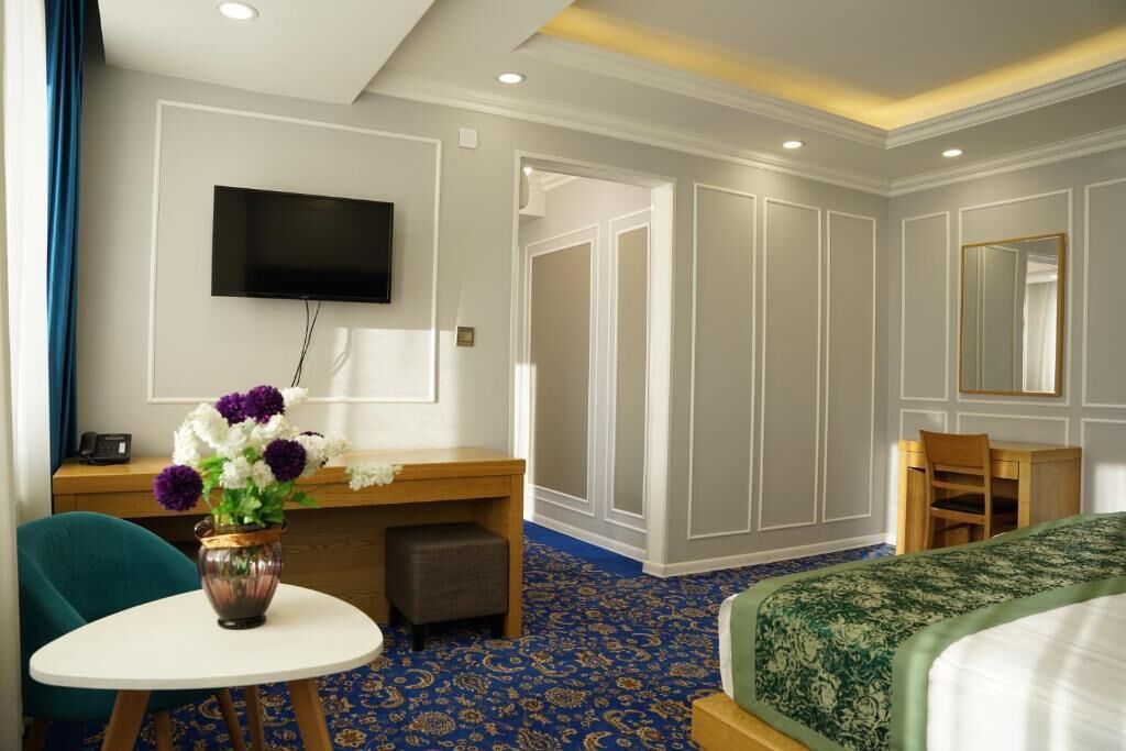 Habitación de hotel con cama, escritorio con silla, televisor montado en la pared y una pequeña mesa con un jarrón de flores.