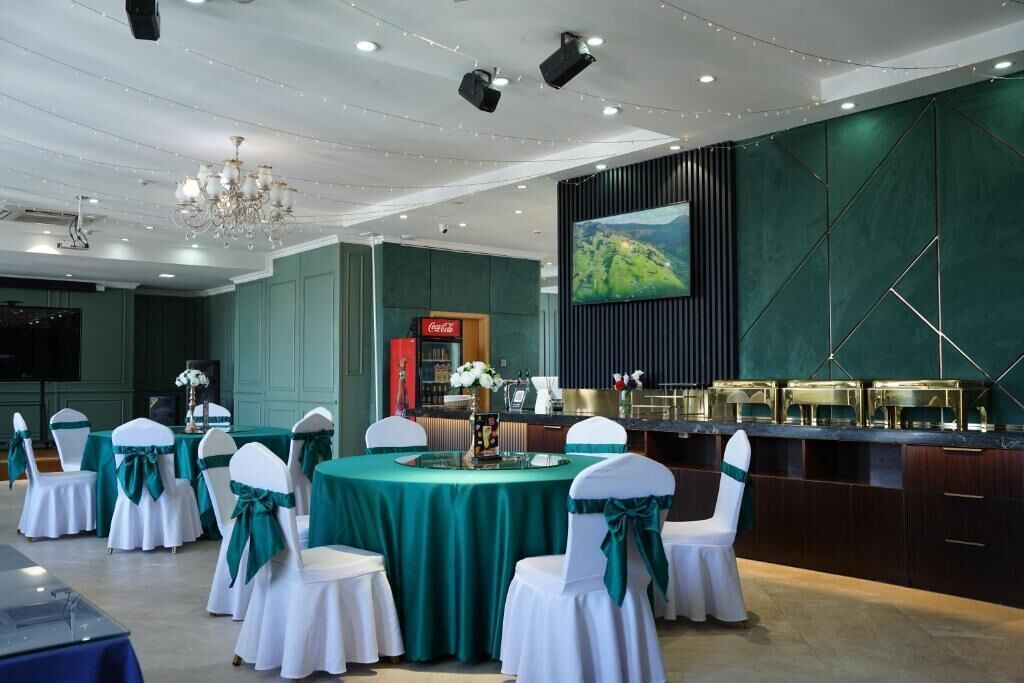 Un salón de banquetes con mesas redondas, sillas con cintas verdes, una zona de bar y una gran pantalla que muestra un paisaje.