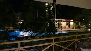 Vue nocturne d’un espace piscine avec une terrasse en bois, des chaises et un bâtiment dont l’entrée est illuminée.