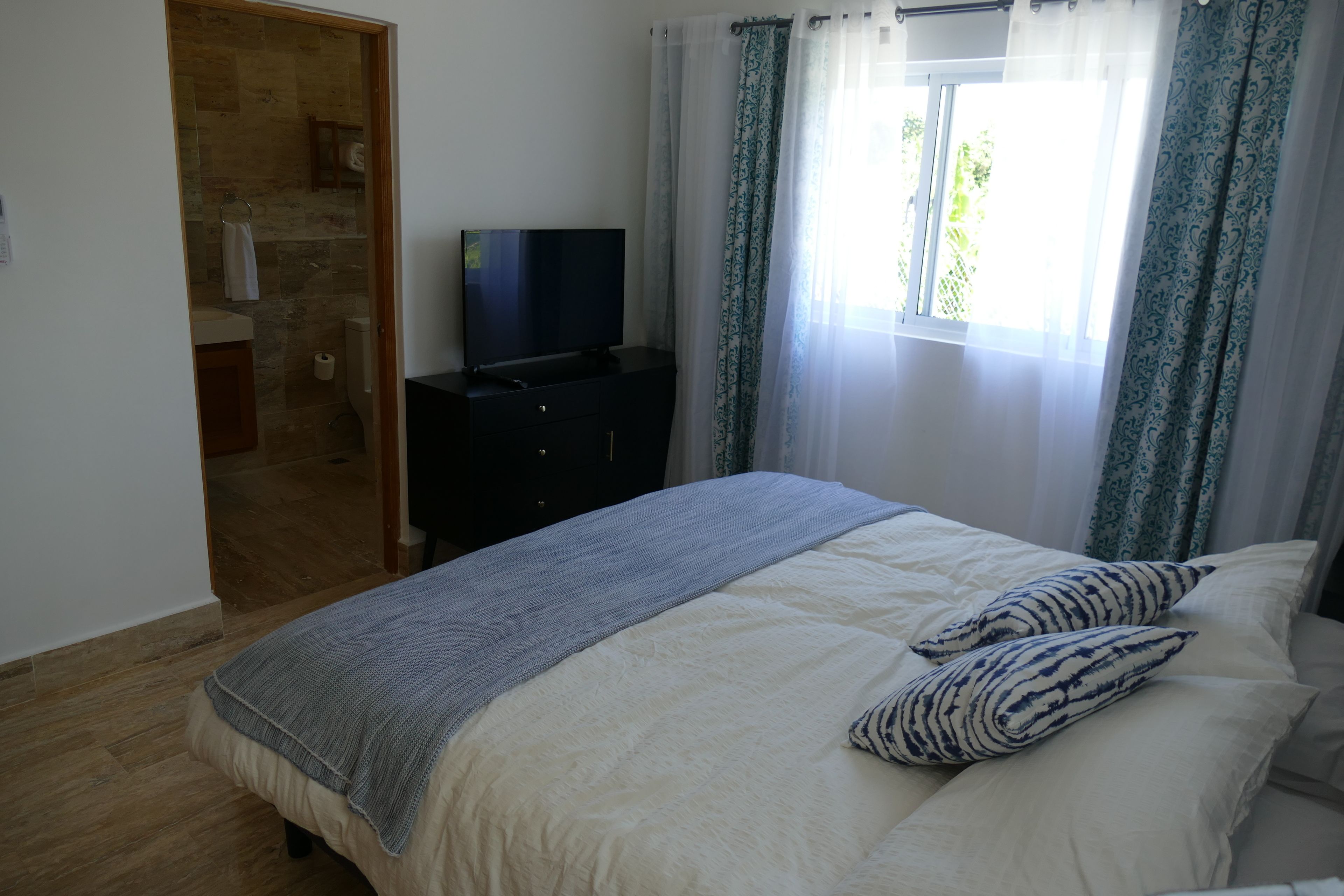 Een slaapkamer met een bed, een tv op een dressoir, een raam met gordijnen en een badkamer zichtbaar door een geopende deur.