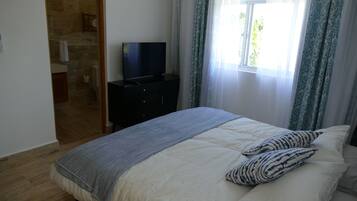 Een slaapkamer met een bed, een tv op een dressoir, een raam met gordijnen en een badkamer zichtbaar door een geopende deur.