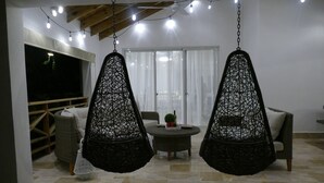 Une pièce avec deux chaises noires suspendues, un téléviseur, une petite table ronde avec une plante et une grande fenêtre avec des rideaux.