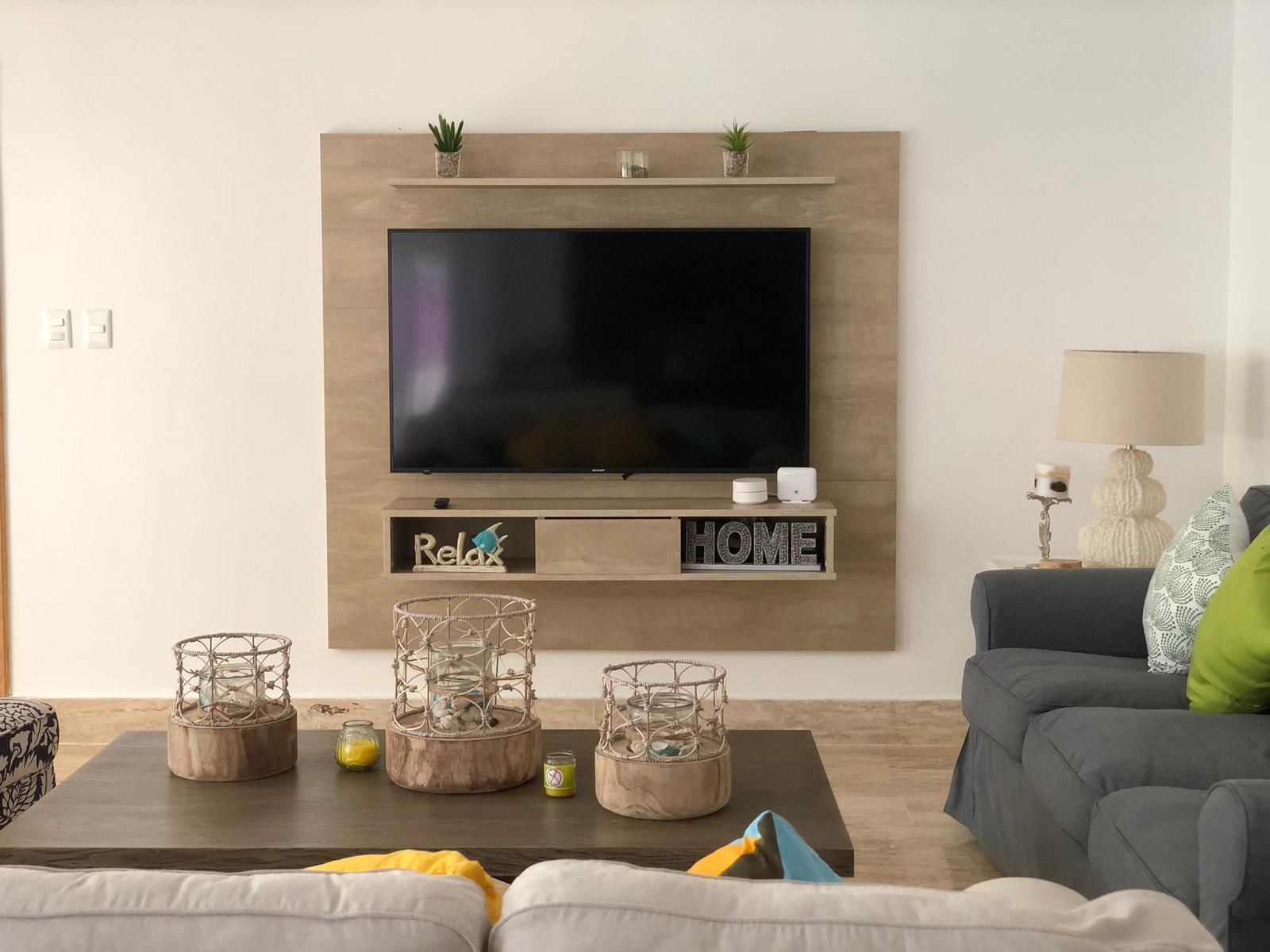 A modern living room with a flat-screen TV mounted on a wooden wall, a sofa, a coffee table with decorative items, and a lamp.