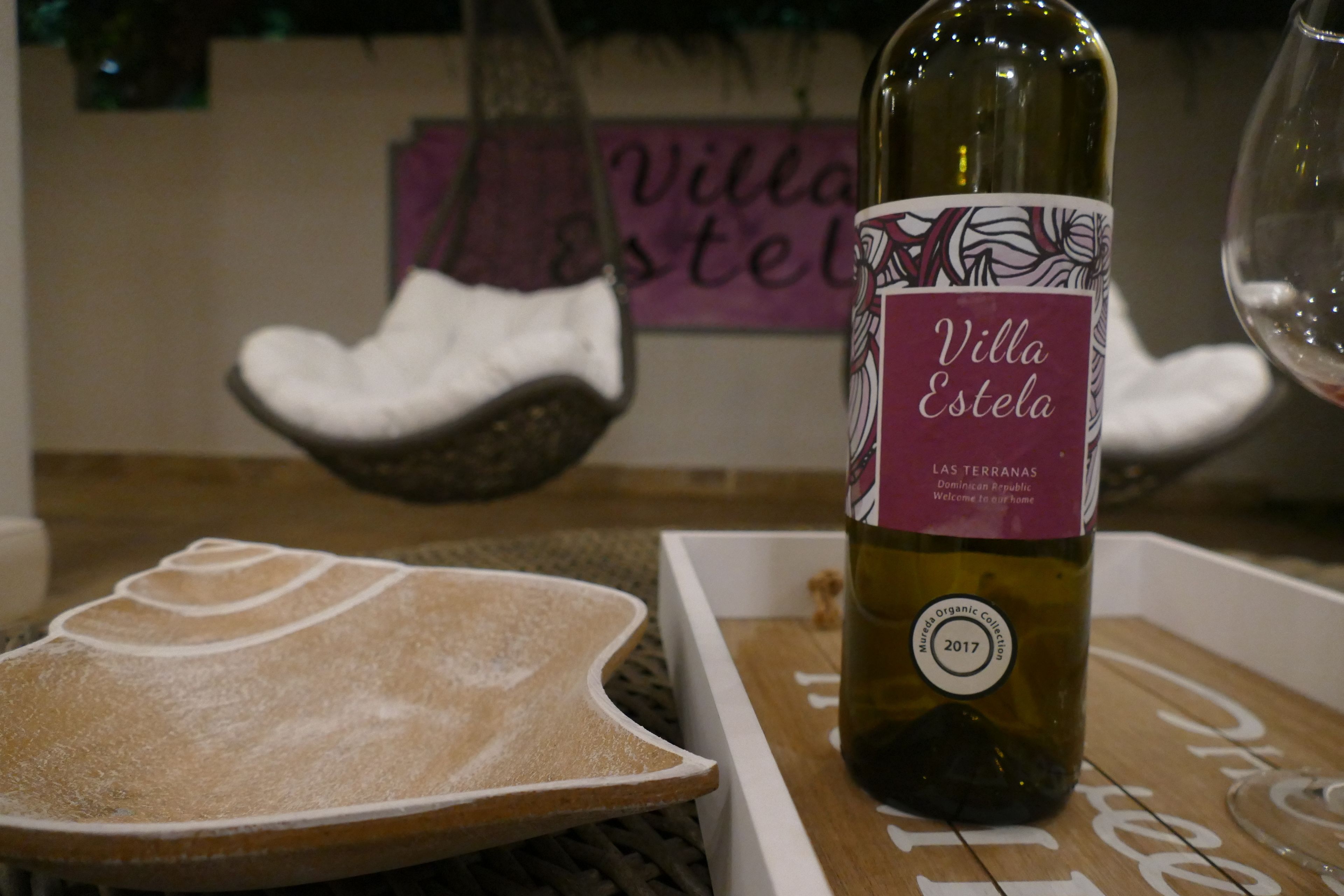 Uma garrafa de vinho Villa Estela, um tabuleiro de madeira com um objeto decorativo e uma cadeira suspensa num quarto com uma placa que diz "Villa Estela".