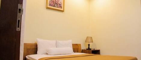 A single bed with white and brown bedding, a nightstand with a lamp, and a framed floral painting on the wall.
