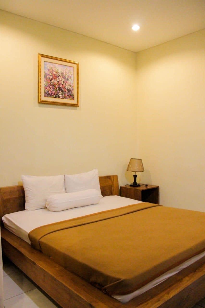 A single bed with a wooden headboard, a bedside table with a lamp, and a framed floral painting on the wall.