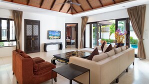 A modern living room with a sofa, a coffee table, and a TV. There is a ceiling fan, a window with curtains, and a view of the outdoors. - Phuket Ratchamaka Pool Villa (Chalong)