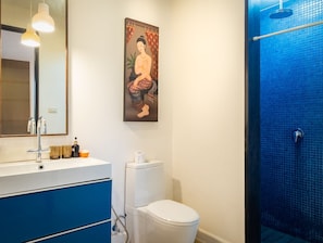 A bathroom with a white toilet, a blue shower, a sink with a mirror, and a painting on the wall.