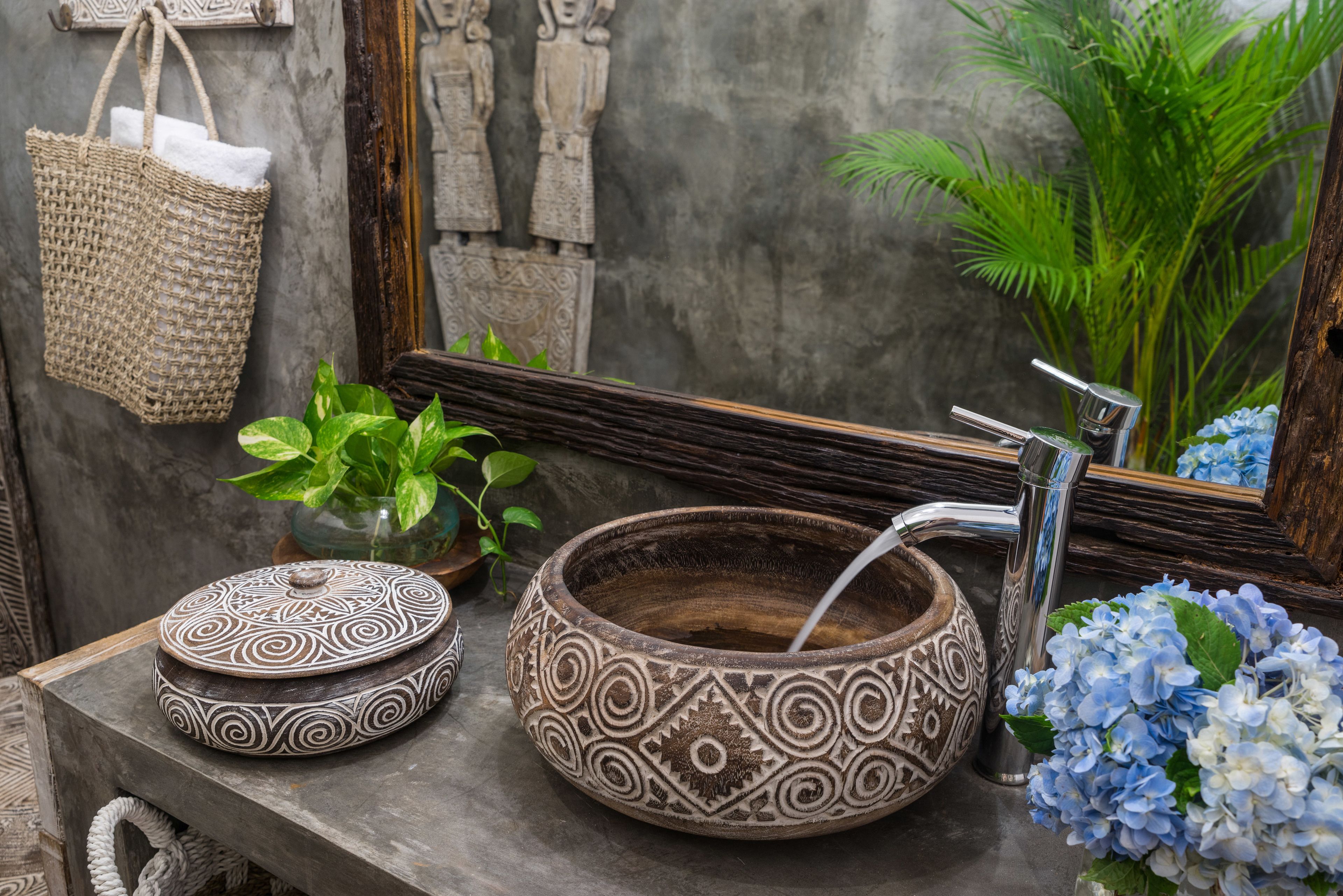 Une salle de bain avec un lavabo en forme de bol en bois, un miroir, un sac tressé et une plante en pot.