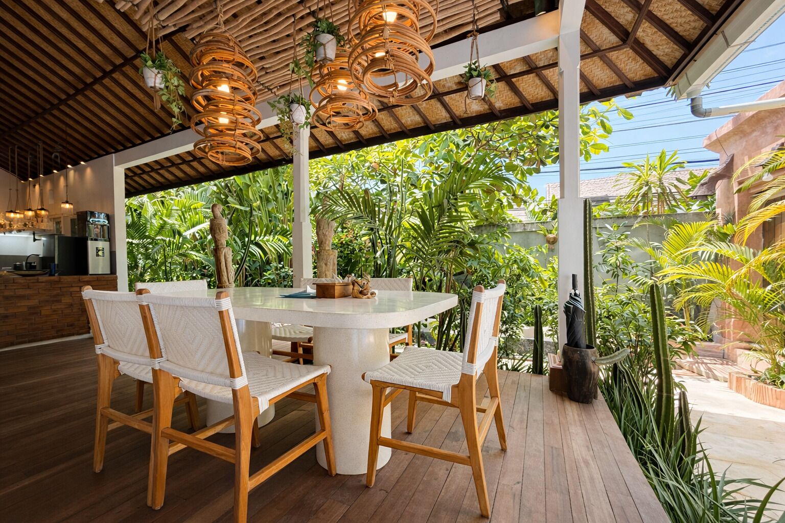 A covered outdoor dining area with wicker chairs and a central table, surrounded by lush greenery.