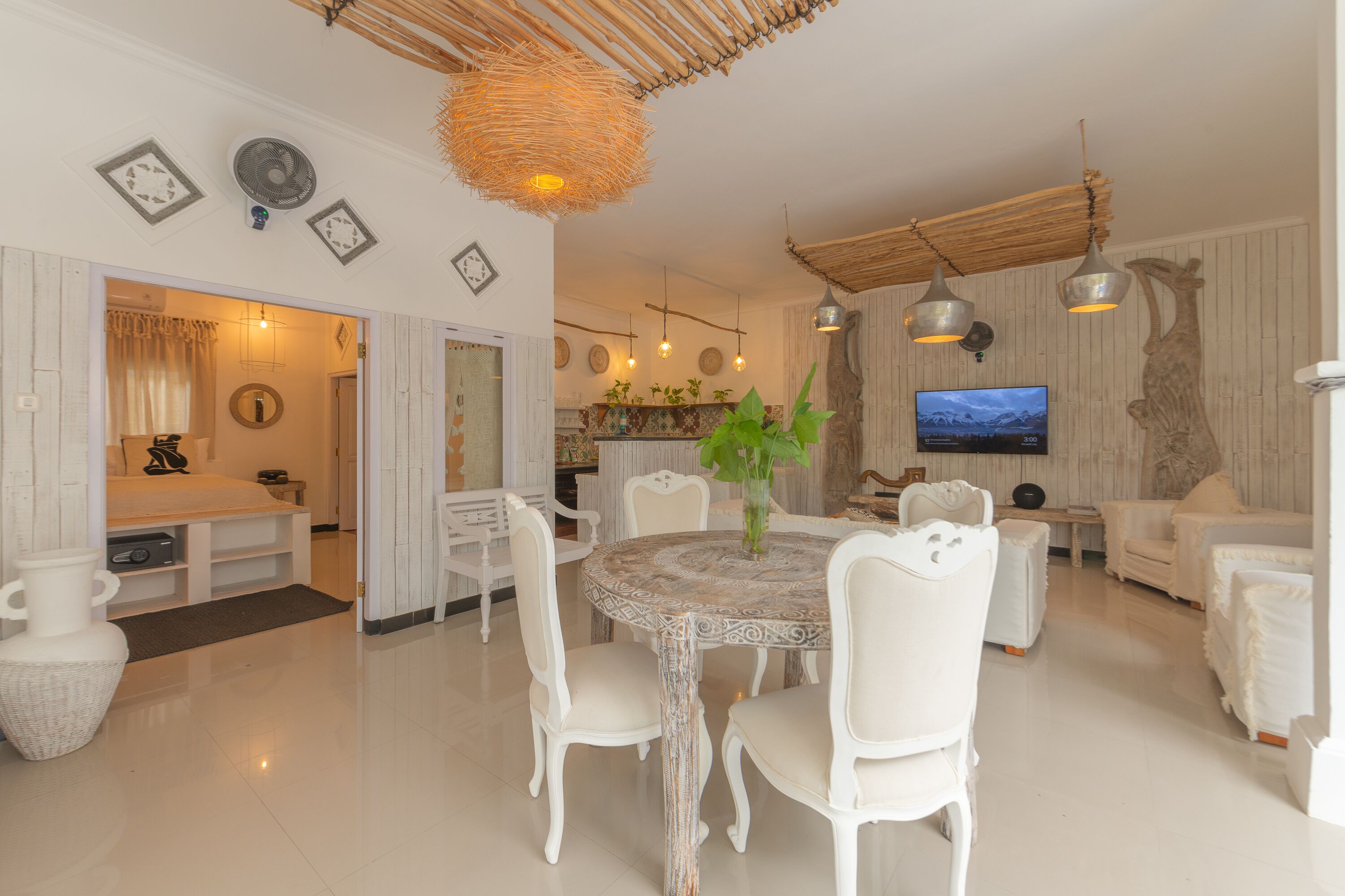 A spacious living area with a round dining table surrounded by white chairs, a large vase with green plants, and a television mounted on the wall.