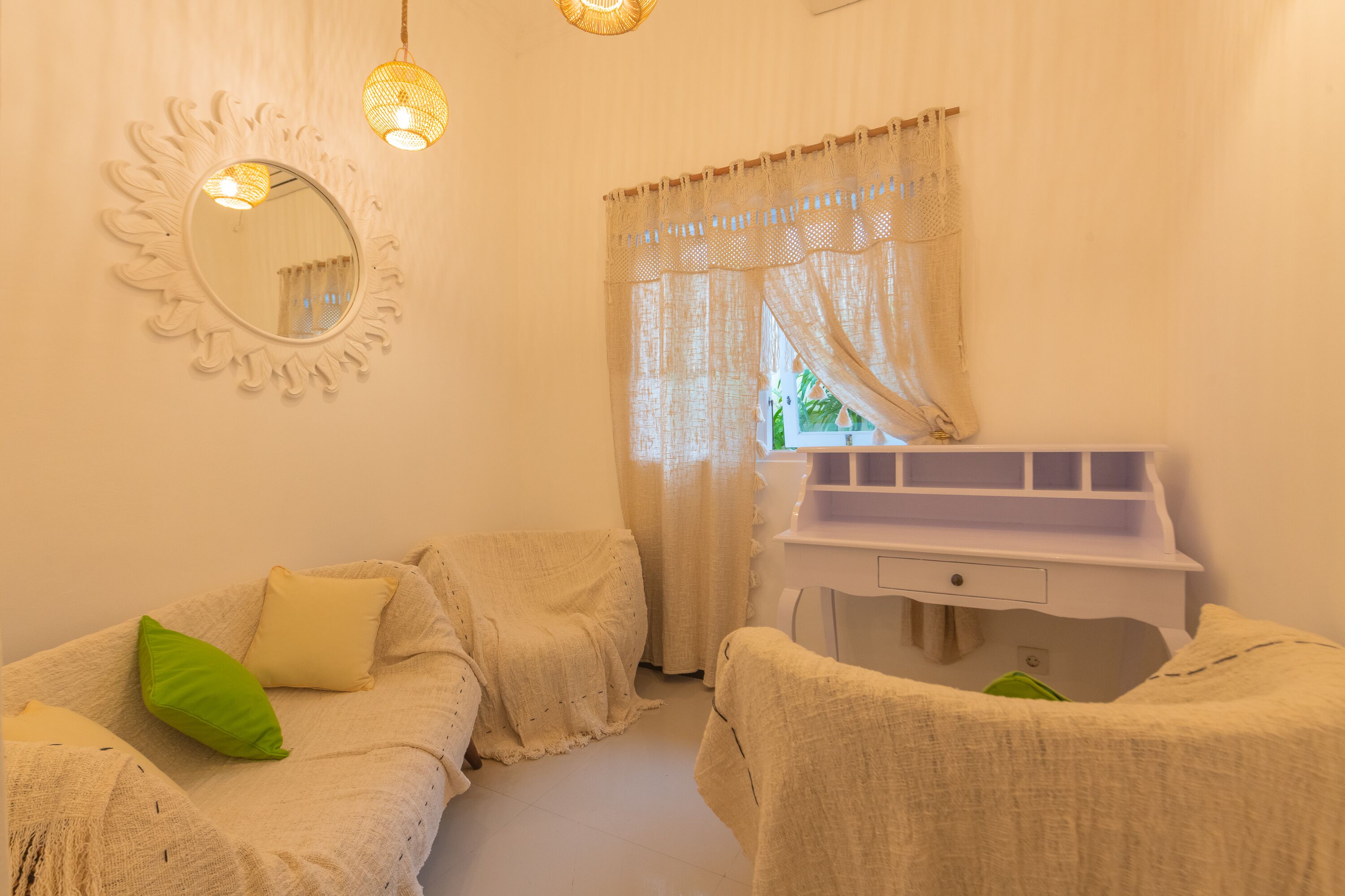 A room with a white vanity, two covered chairs, a mirror with a sunburst design, and a window with sheer curtains.