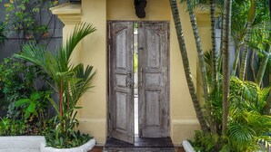A wooden door with intricate carvings, flanked by white planters with tropical plants, leading to a building with a yellow facade. - Smiling Cactus Villa (Seminyak)