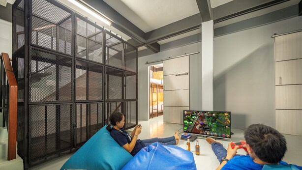 Een kamer met een groot scherm waarop een videospel wordt afgespeeld, iemand die het spel speelt, en een ander persoon zittend op een relaxfauteuil.