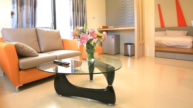 A modern living room with a glass coffee table, a sofa, a vase with flowers, and a bed with a grey headboard. - Amethyst Bay (Magong)
