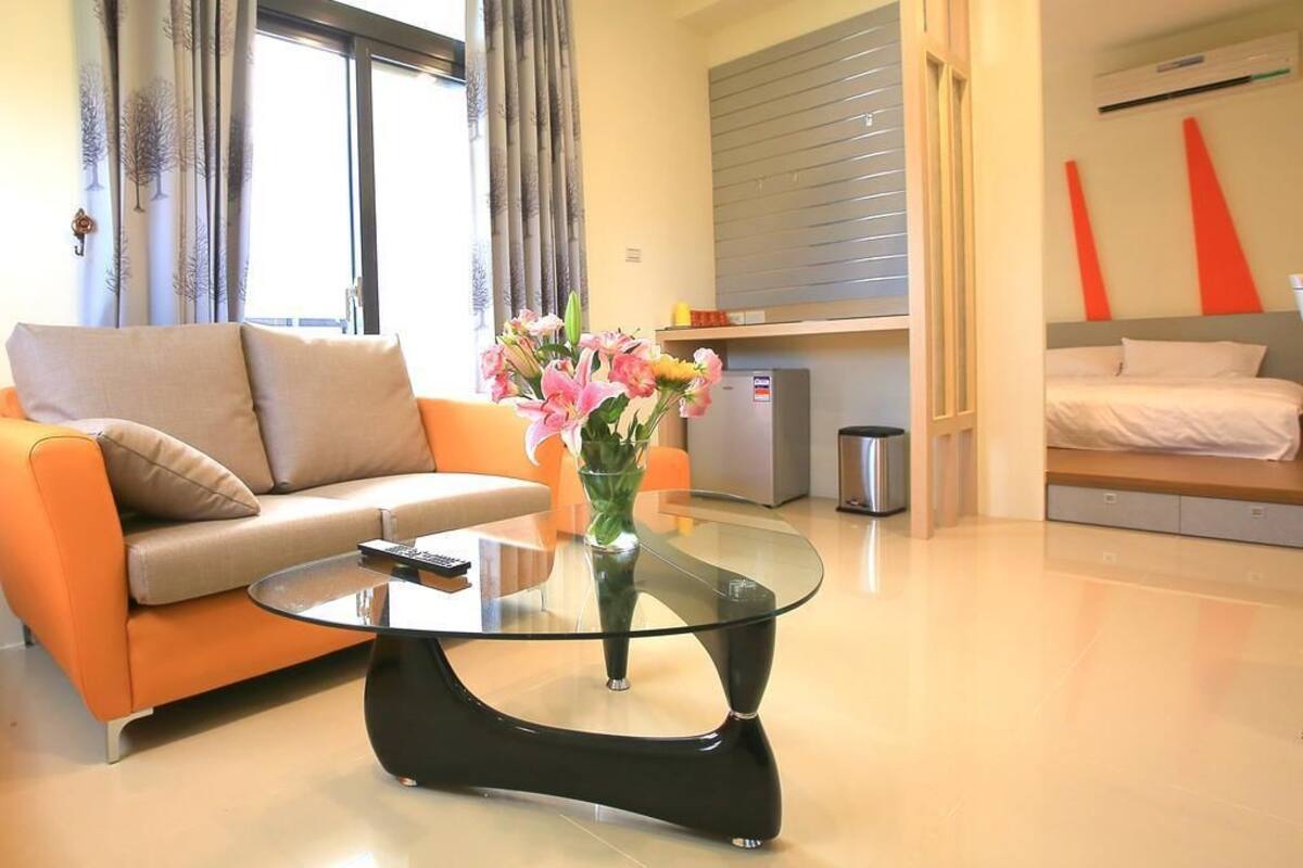 A modern living room with a glass coffee table, a sofa, a vase with flowers, and a bed with a grey headboard.