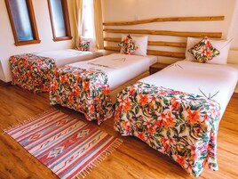 Habitación de hotel con tres camas individuales, cada una con una colcha estampada y ropa de cama blanca. Tiene un cabecero de madera, una ventana con cortinas y una alfombra a rayas sobre el piso de madera.