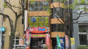 A multi-story building with a brick facade and large windows. There are several banners and signs in front of the entrance, indicating various businesses. - AMP FLAT Nishijin5 (Fukuoka)